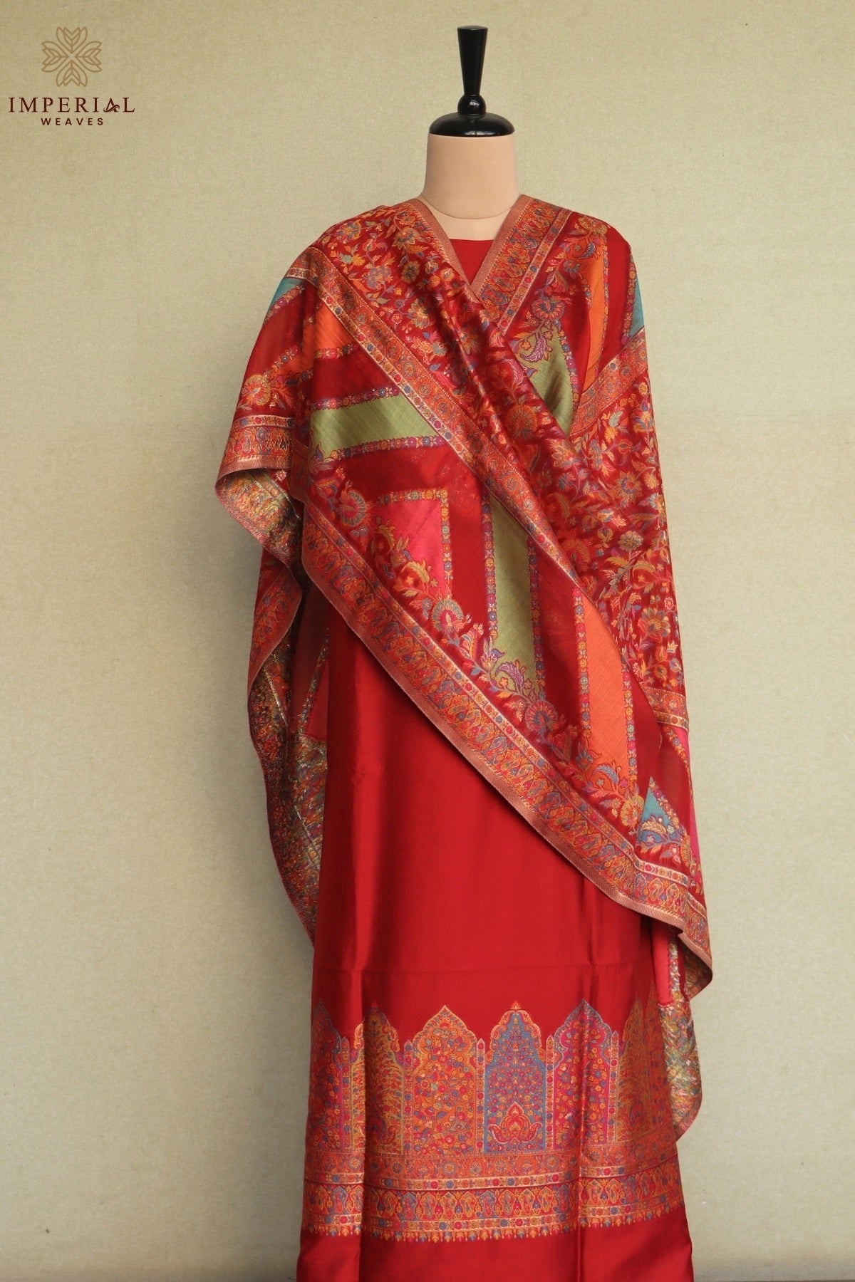 A red traditional outfit displayed on a mannequin against a light beige wall, featuring a draped shawl with intricate multicolor patterns and a matching patterned border along the bottom hem. Kani Suit 