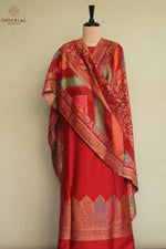 A red traditional outfit displayed on a mannequin against a light beige wall, featuring a draped shawl with intricate multicolor patterns and a matching patterned border along the bottom hem. Kani Suit 