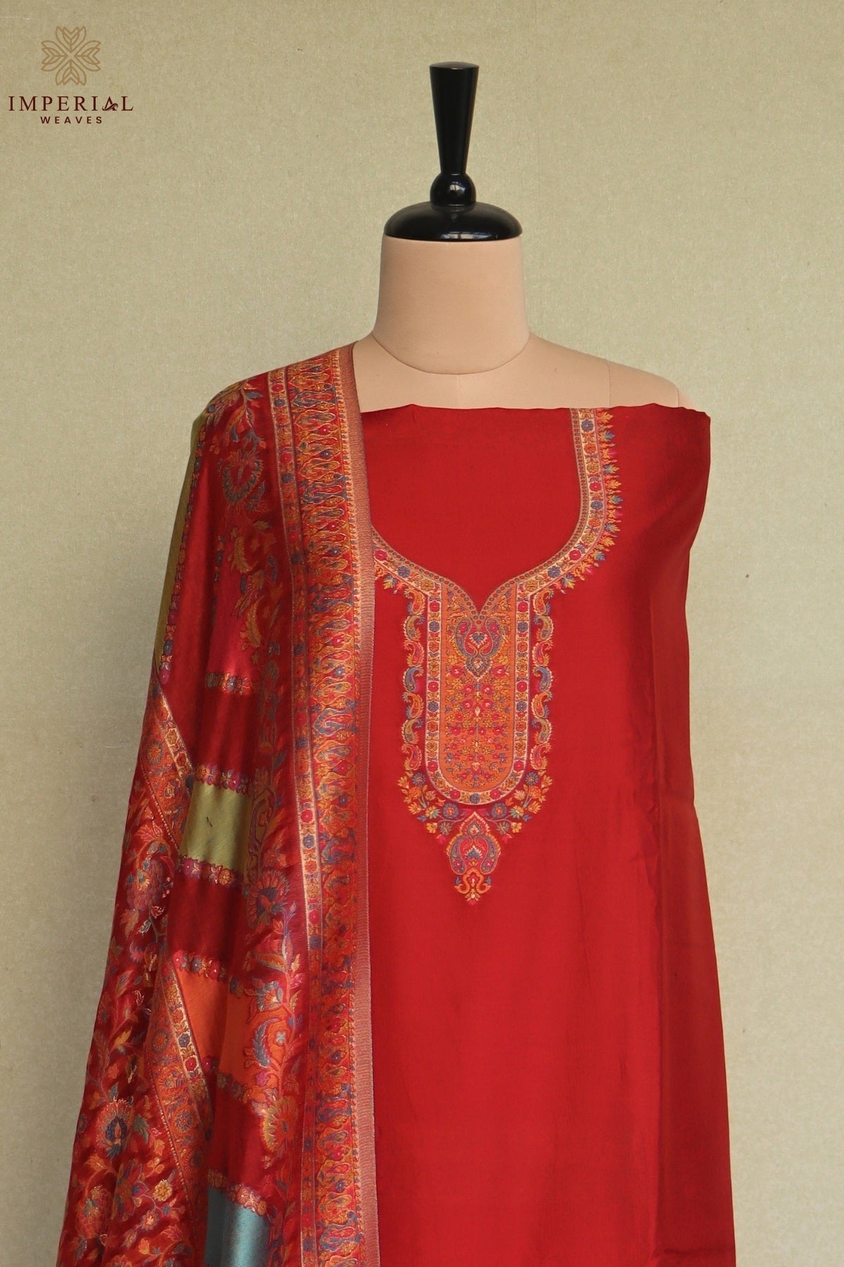A red women's salwar suit set displayed on a mannequin, featuring a solid red kurta with ornate multicolor embroidery around the neckline and a matching patterned dupatta draped over the shoulder. Kashmiri Kani Silk Suit